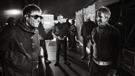 Liam Gallagher and Noel Gallagher share a rare, smiling moment backstage in a grainy black-and-white photo while surrounded by their crew.