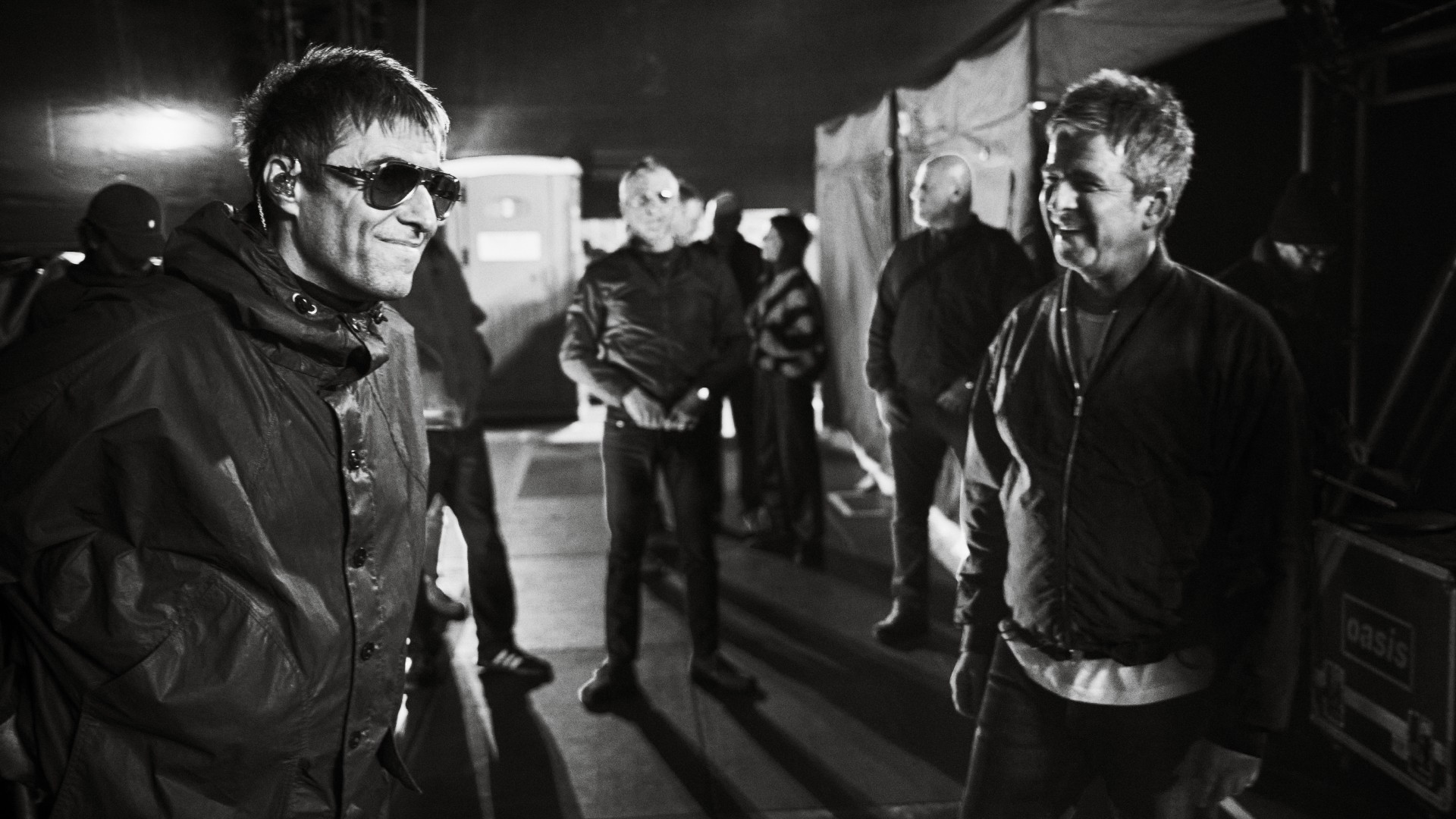 Liam Gallagher and Noel Gallagher share a rare, smiling moment backstage in a grainy black-and-white photo while surrounded by their crew.