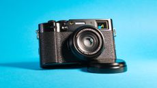 The Fujifilm X100VI mirrorless camera against a blue background propped up using the lens cap.