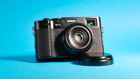 The Fujifilm X100VI mirrorless camera against a blue background propped up using the lens cap.