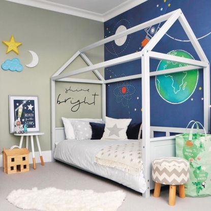 Child's bedroom with four poster bed, mural of planets, grey carpet, chest of drawers and bedding.