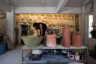 A young woman wearing a black, sleeveless T-shirt and trousers works on some cement planters in her dimly lit industrial studio.