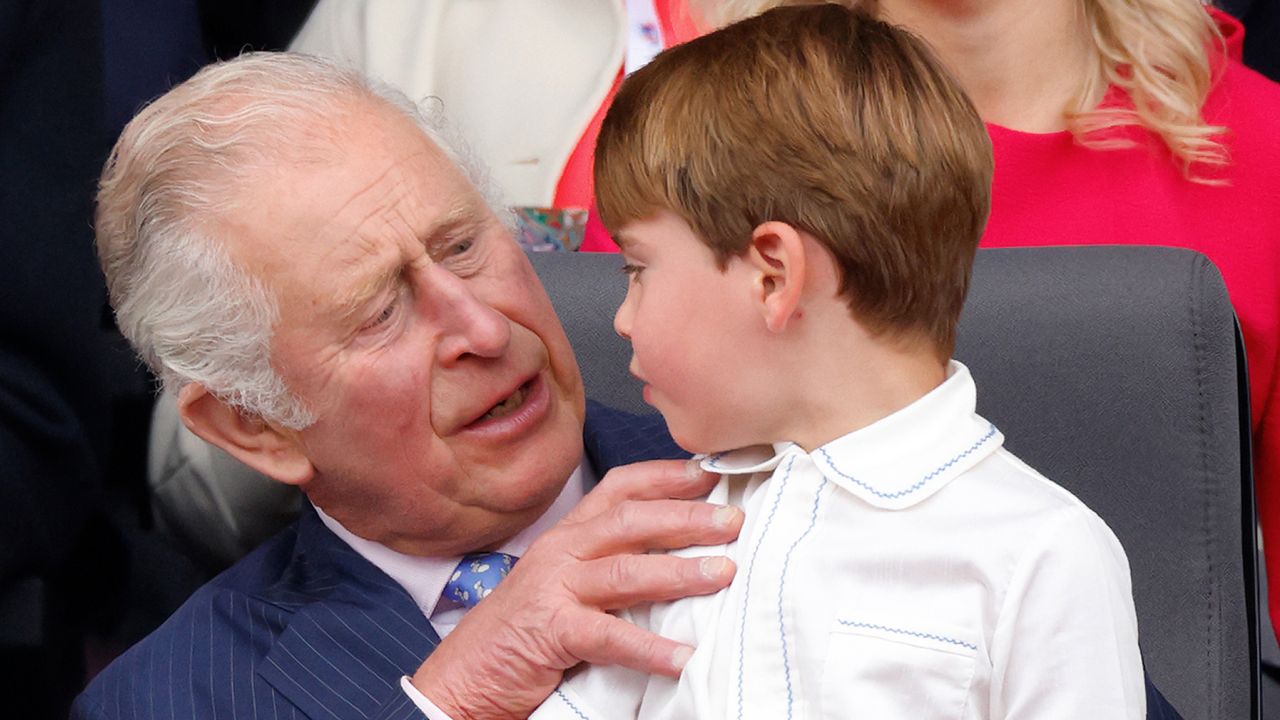 King Charles sitting in a chair wearing a blue suit and a blue tie with Prince Louis, dressed in a white shirt, on his lap 