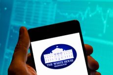 Person holding smartphone with White House logo on it and stock chart blurred in the background