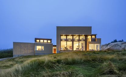 Exterior view of Float House in the evening with the lights on. The house sits on a rocky and grassy terrain