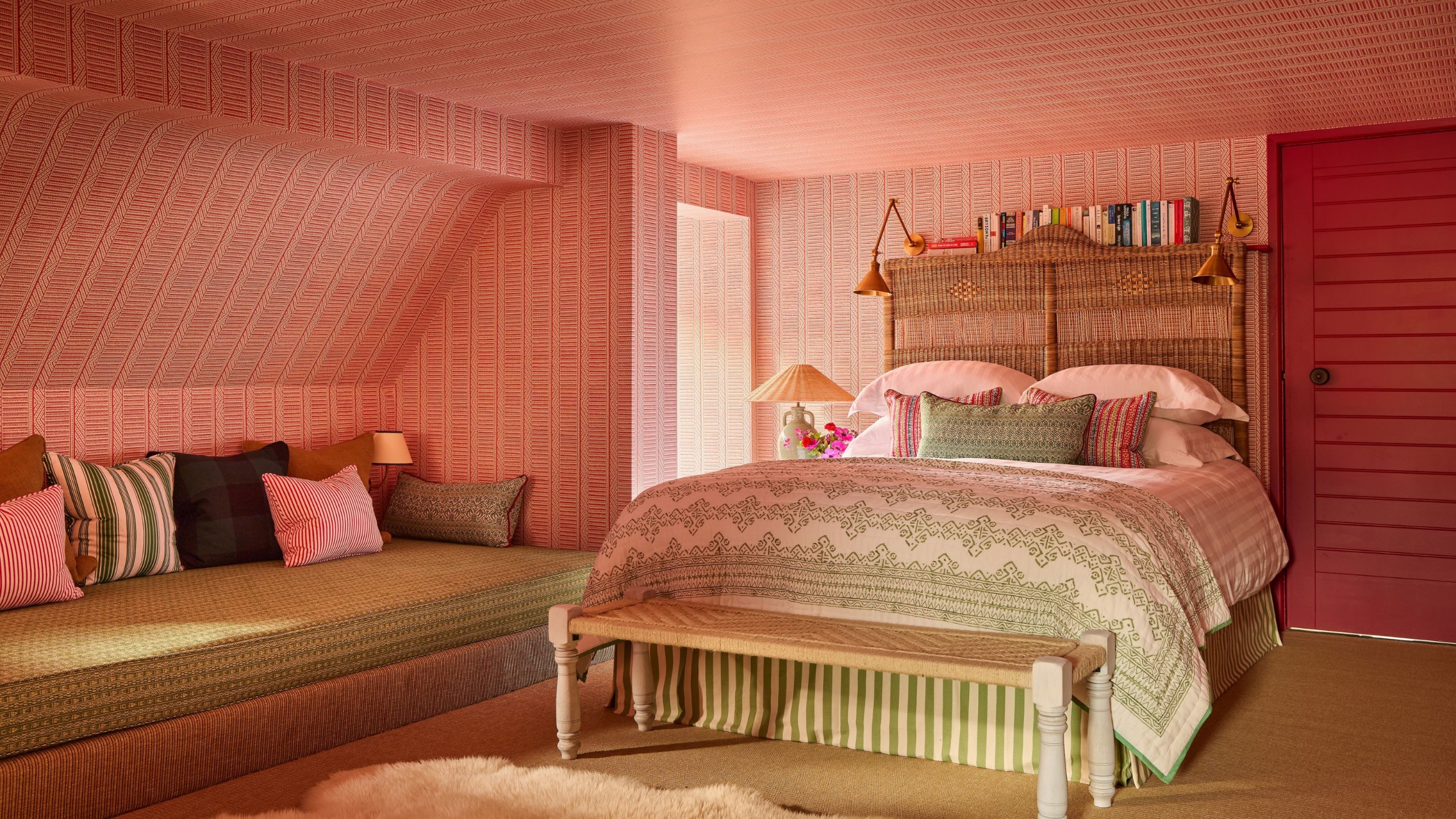 An attic bedroom with a large bed with a cane headboard, sage green patterned bedding with raspberry red cushions, and a warm pink patterned wallpaper all over the walls and ceiling, with a seating nook in the eaves.