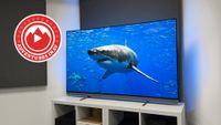 The 65-inch Philips OLED910 TV photographed on a white, wooden stand. On the screen is an image of a great white shark, and in the corner of the photo is a red logo that says 'Adventures In AV'.