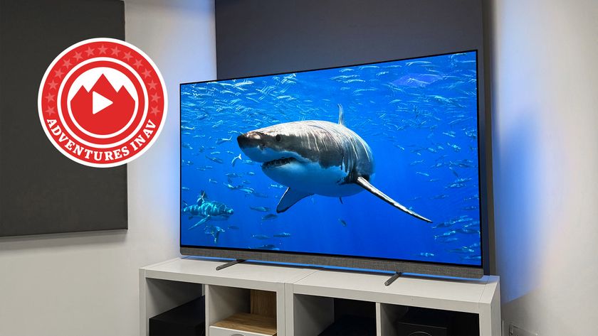 The 65-inch Philips OLED910 TV photographed on a white, wooden stand. On the screen is an image of a great white shark, and in the corner of the photo is a red logo that says &#039;Adventures In AV&#039;.