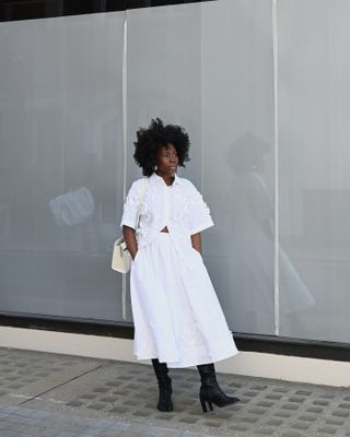 Danielle Jinadu mengenakan kemeja putih berkancing dan rok putih A-line dari COS dengan sepatu hitam dan tas putih.