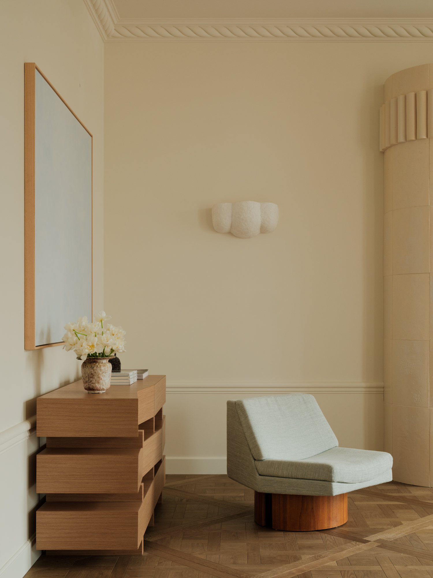 Image of the corner of a luxury, but minimal living room. There are wooden floors and warm, off-white walls. There is a wooden console table and a light blue, modern geometric chair.