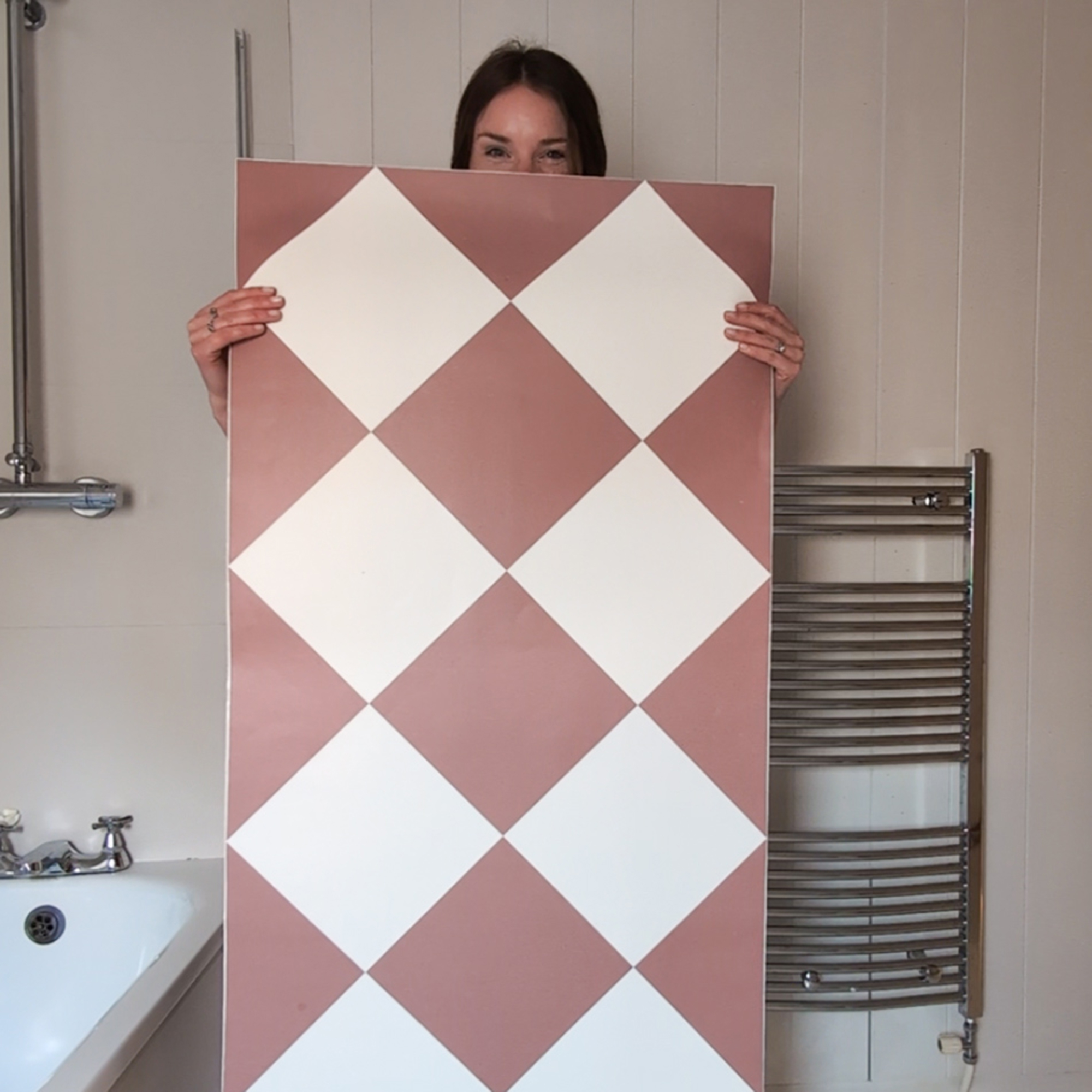 Lara Winter standing in a beige bathroom holding a panel of vinyl floor tiles with a terracotta and white checkerboard design
