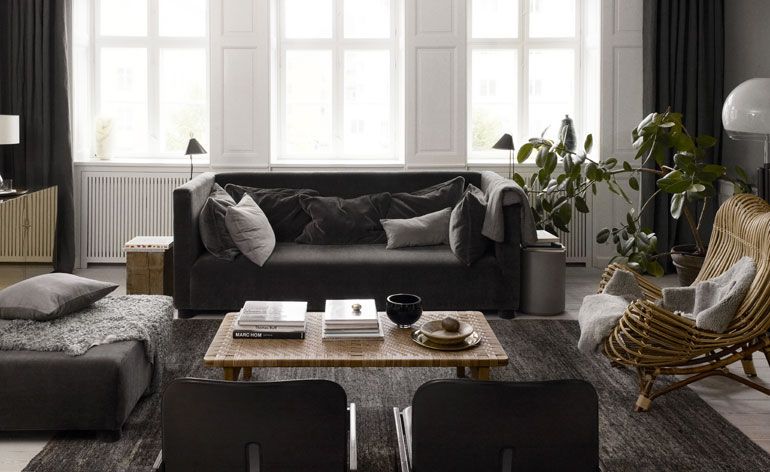 Interior view of the living area at The Apartment gallery featuring grey seating with cushions, a coffee table with books and other items on top, a rug, a green plant in a pot, a natural chair and windows with curtains pulled to the sides