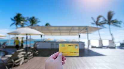 A hand holds a credit card in the foreground while a beach resort bar is in the background