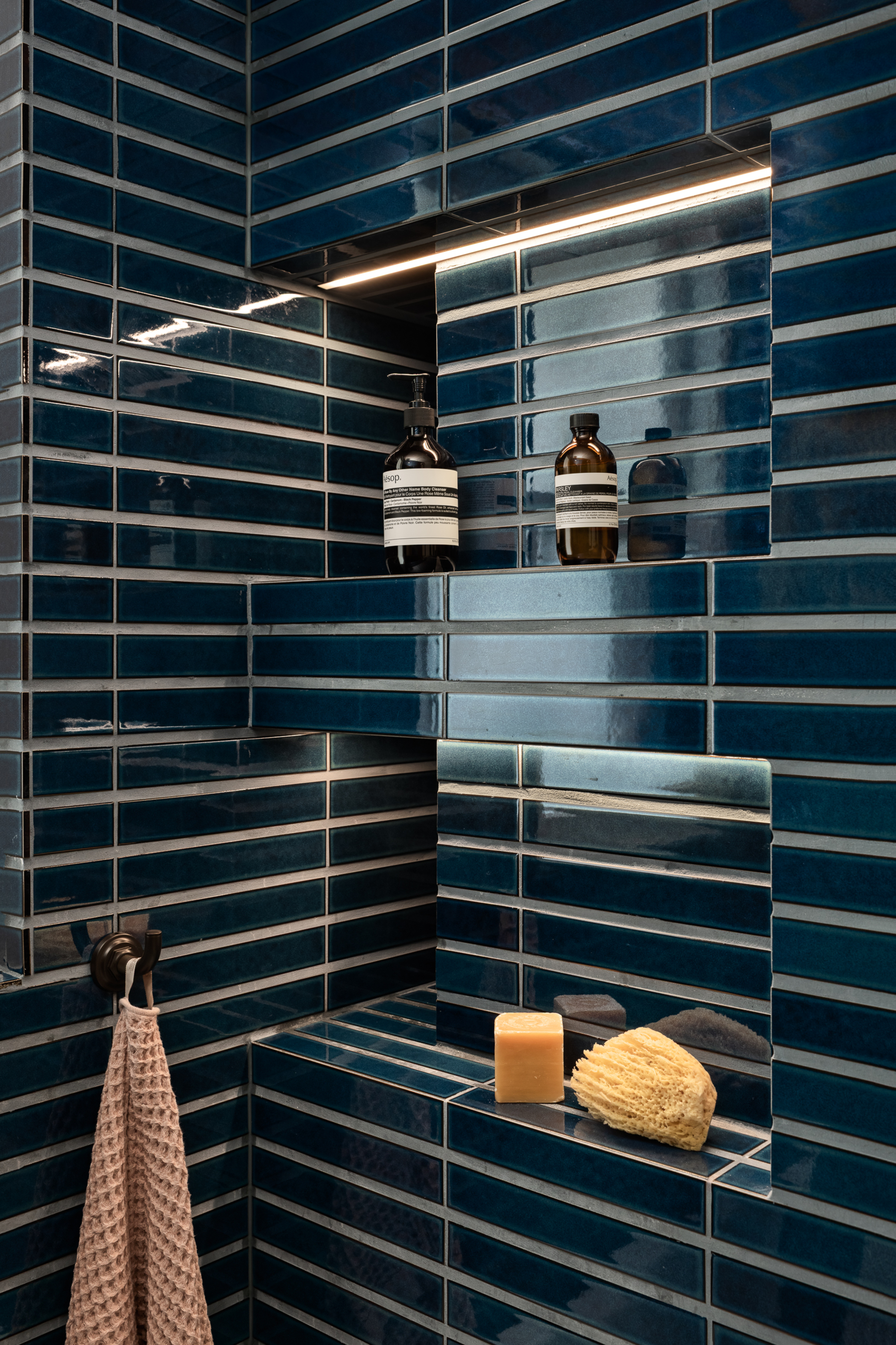 blue tiles in shower with shower niches with LED strips, loofah and soap, and aesop body products with a towel on a hook