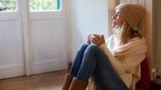 Woman sat next to a radiator wearing a shawl and hat, drinking a hot drink 