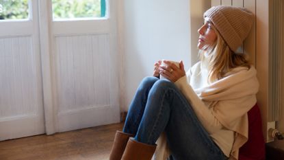 Woman sat next to a radiator wearing a shawl and hat, drinking a hot drink