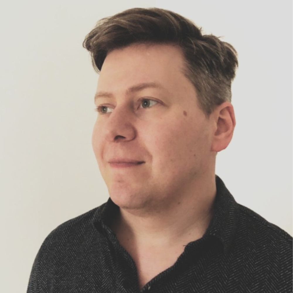 Man with short brown hair, wearing a dark herringbone shirt, looking to the side against a plain light background.