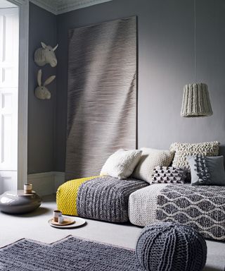 Grey living room ideas featuring texture in the form of chunky knit rugs, throws and a pouffe, a pale grey wall hanging and textured cushions.