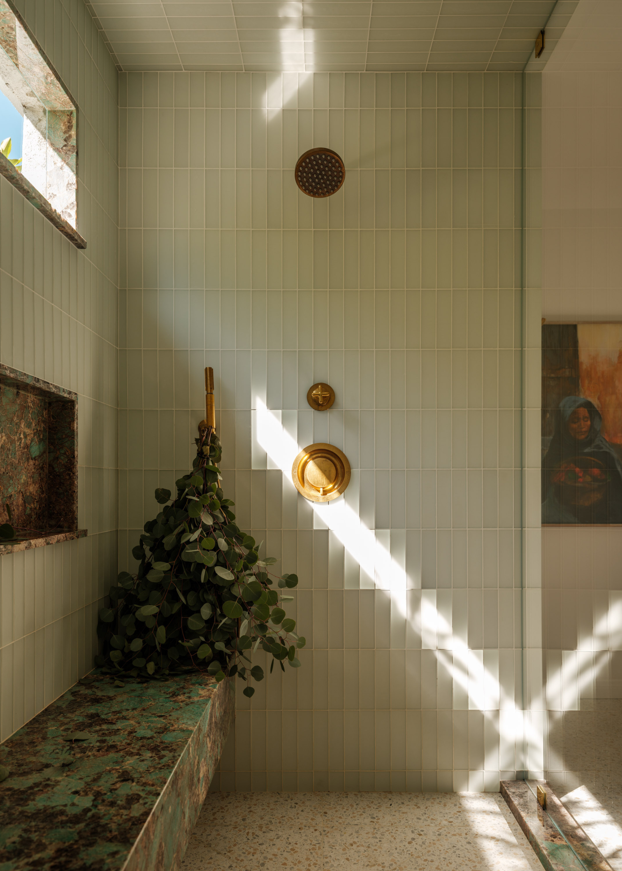 A shower room with pale glassy tiles, brass hardware, a blue marble shower bench, and a bundle of eucalyptus