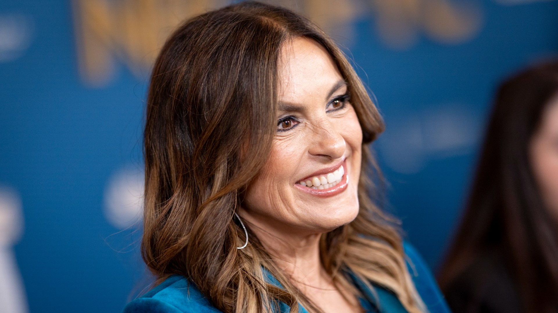 a close-up photo of a white woman with brown hair smiling on a red carpet in front of a blue backdrop