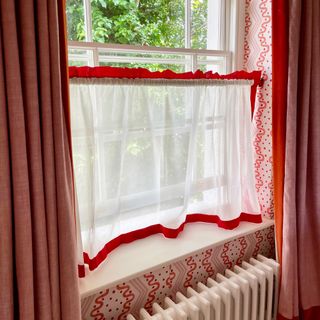 Red trim cafe curtain in living room with pink curtains and wallpaper.