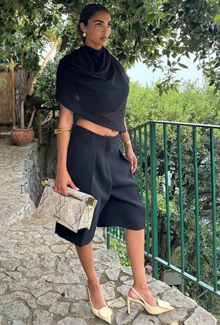 a woman's outfit showing what to wear in 80-degree weather with a black draped top with black bermuda shorts styled with yellow slingback heels and a snakeskin clutch bag
