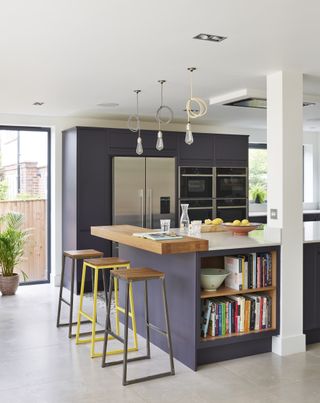 A purple kitchen with tiled floor, bar stools, kitchen island and a wooden breakfast bar with colorful books underneath