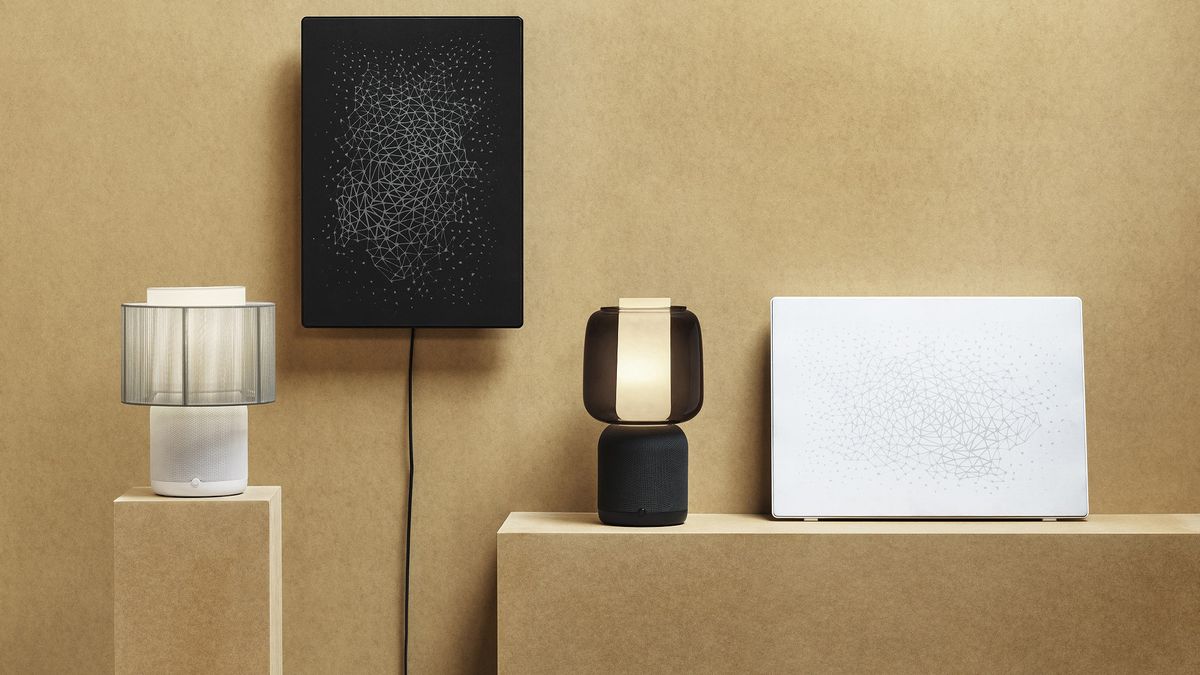 The IKEA Symfonisk smart table lamp on a shelf next to other Ikea smart home kit. The shelf and wall behind it are beige. 