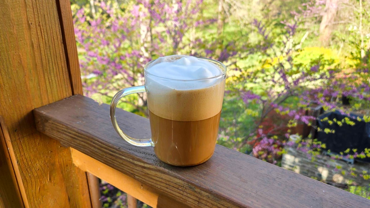 A homemade cappucino rests on the railing of a deck overlooking a garden.
