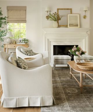 neutral farmhouse living room with a large rug and two skirted armchairs sat on top beside a white wooden fireplace