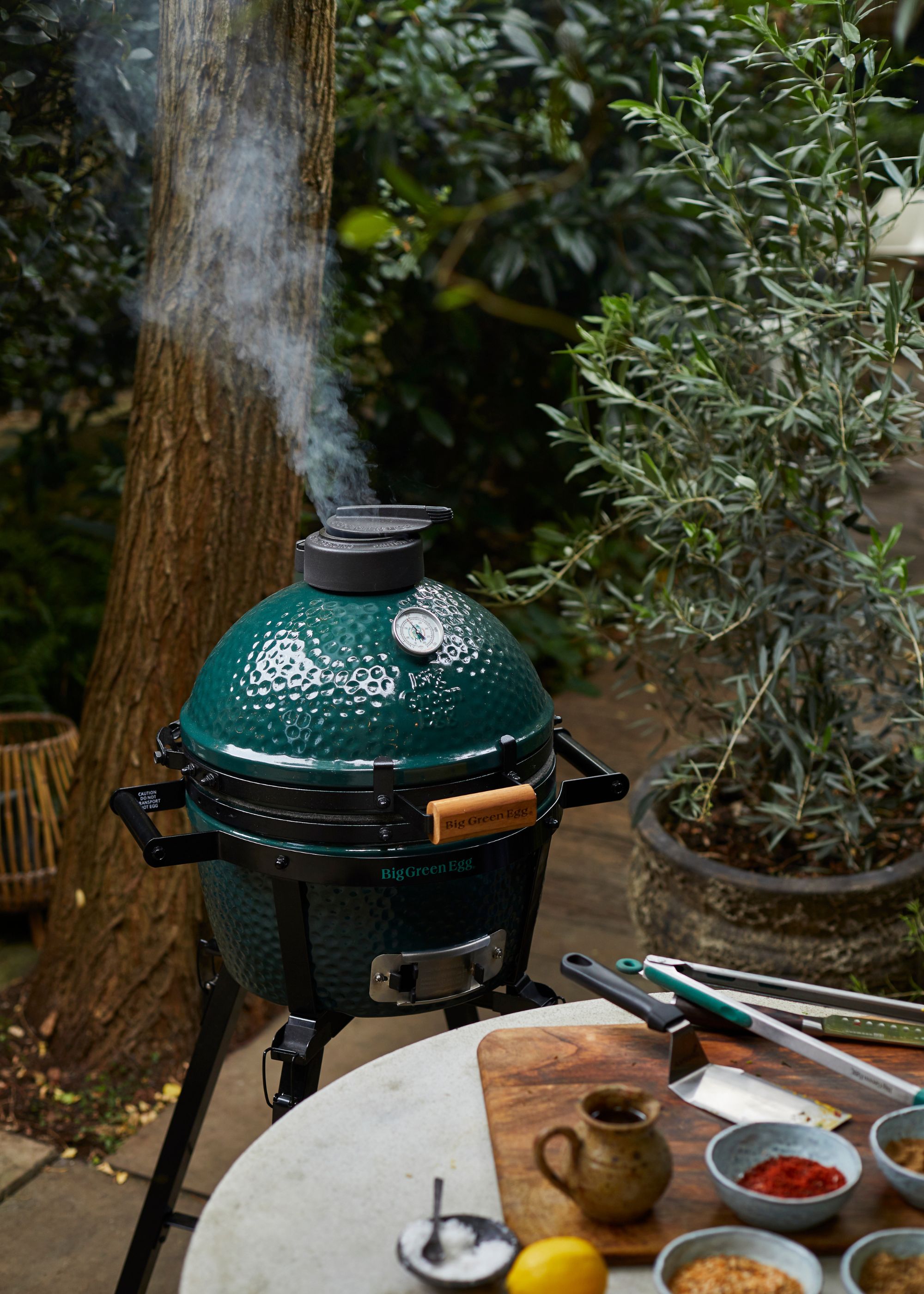 A Big Green Egg with steam coming out of it beside a table of spices and cooking accessories in a backyard