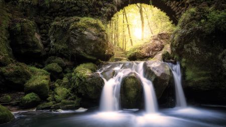 A serene waterfall flows over moss-covered rocks under an ancient stone arch. Sunlight filters through dense forest