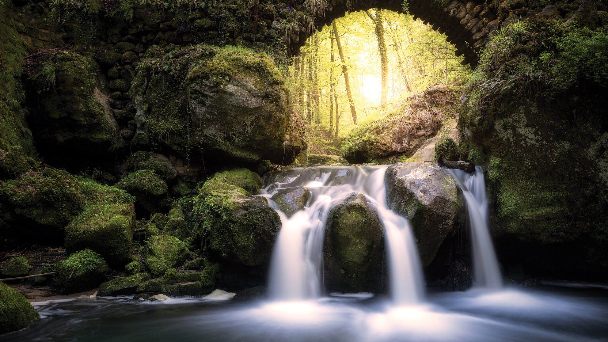 A serene waterfall flows over moss-covered rocks under an ancient stone arch. Sunlight filters through dense forest