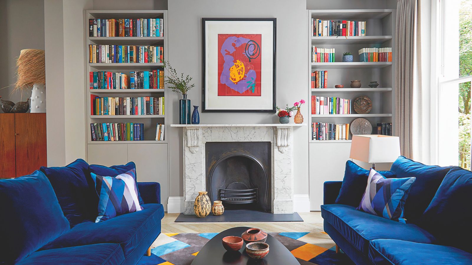 A living room with two blue velvet sofas either side of a a marble surround fireplace. Symmetrical bookshelves on either side, behind. 
