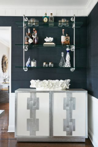 Corner of a dark black-papered room with a white and silver embellished cabinet and wall-hung shelving for bottles and trinkets