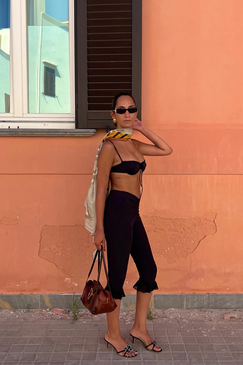 European vacation outfit idea is shown in a photo of a woman standing outside wearing black rectangular sunglasses with gold earrings, a yellow scarf, a black bikini top, black capri pants, black heeled mules, and a red handbag