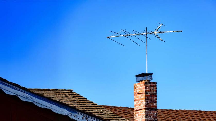 A traditional rooftop TV antenna mounted on a brick chimney rises above the brown shingle roof of a house