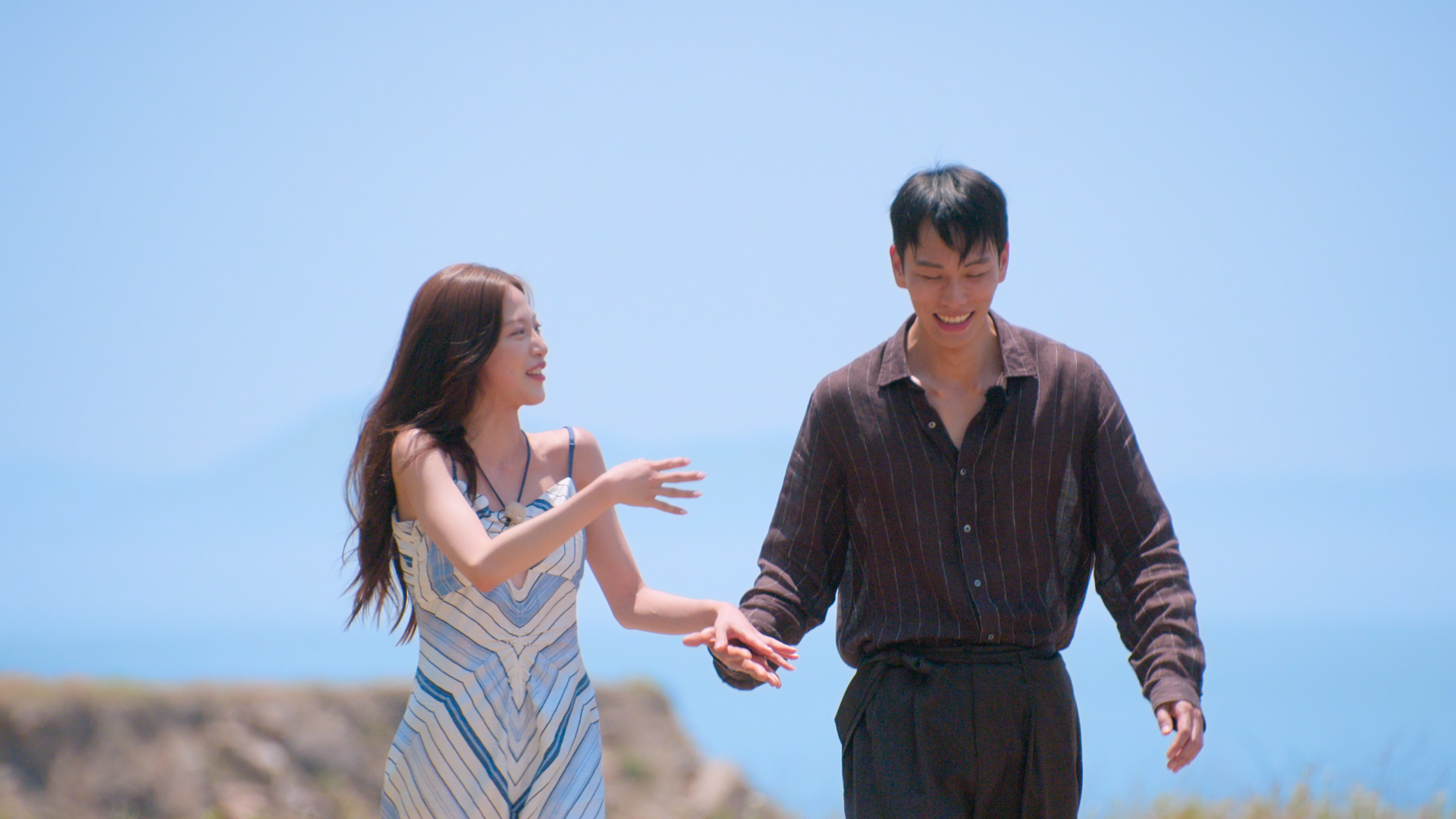 A woman in a geometric-print dress (Lee Joo-young) and a man in a dark shirt and pants (Kim Jae-jin) hold hands and laugh while walking outdoors, with a rocky cliff and the sea behind them, in 'Single's Inferno' season 5.