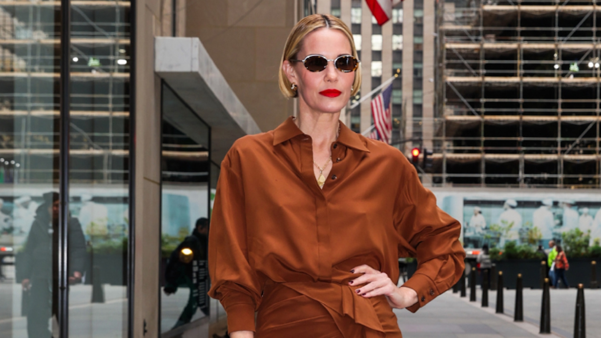 Leslie Bibb wearing a bob hairstyle and a monochromatic tan outfit. She is wearing oval sunglasses and a red lip. 