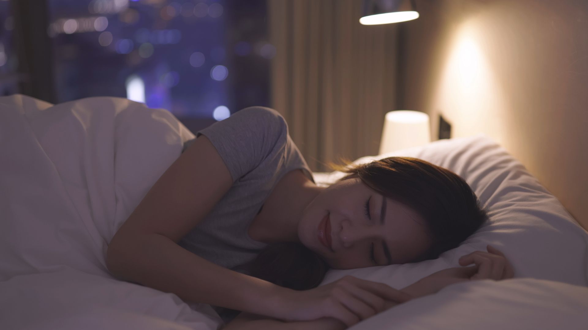 A woman lies in bed on her side at night, appearing to have just fallen asleep.