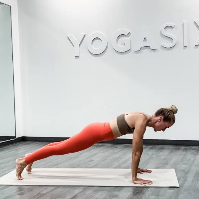 Woman demonstrates yoga pose in yoga studio with words Yoga Six embossed on wall behind her