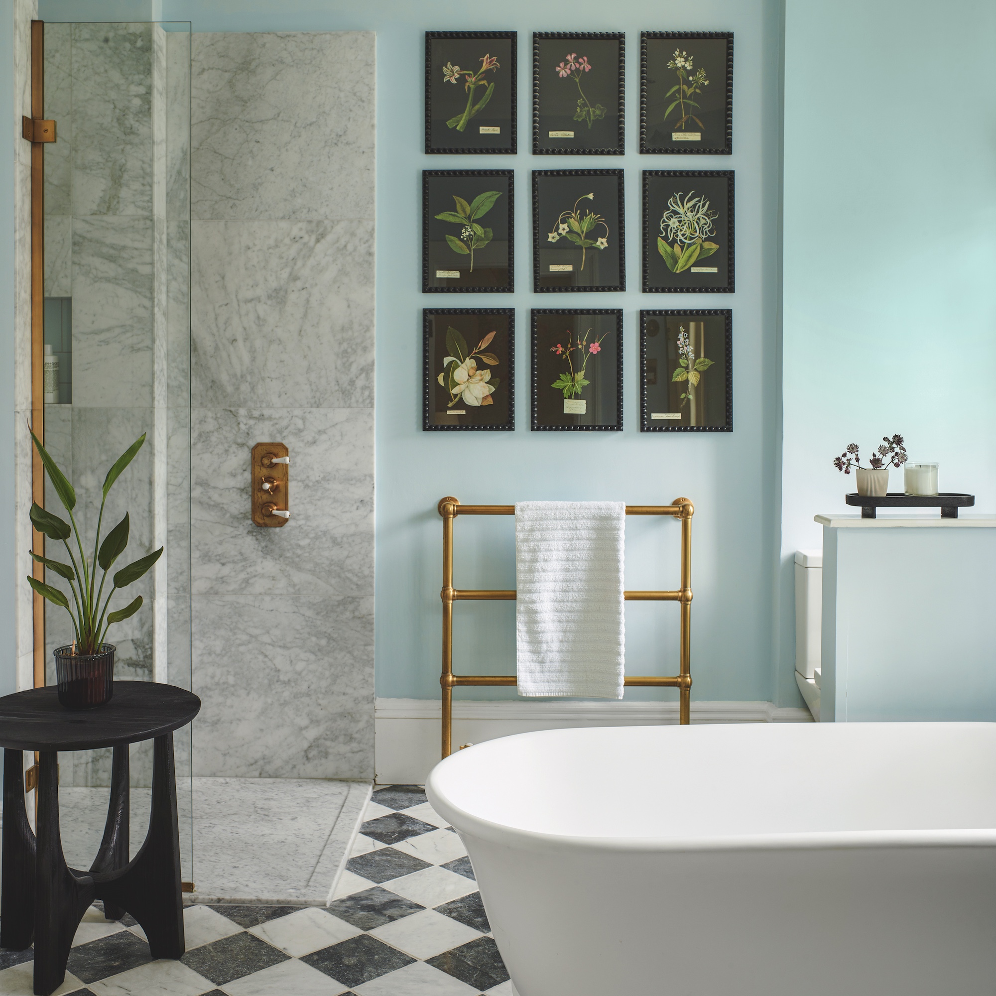 bathroom with white freestanding bath, marble shower enclosure, brass fittings and gallery wall of flower prints with black and white chequerboard floor
