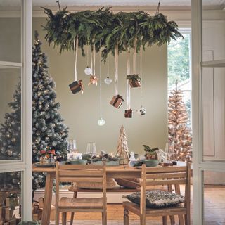 A wooden dining table and chairs in a festive dining room. Presents and baubles hang from above and the table is decorated with ornaments and glasses