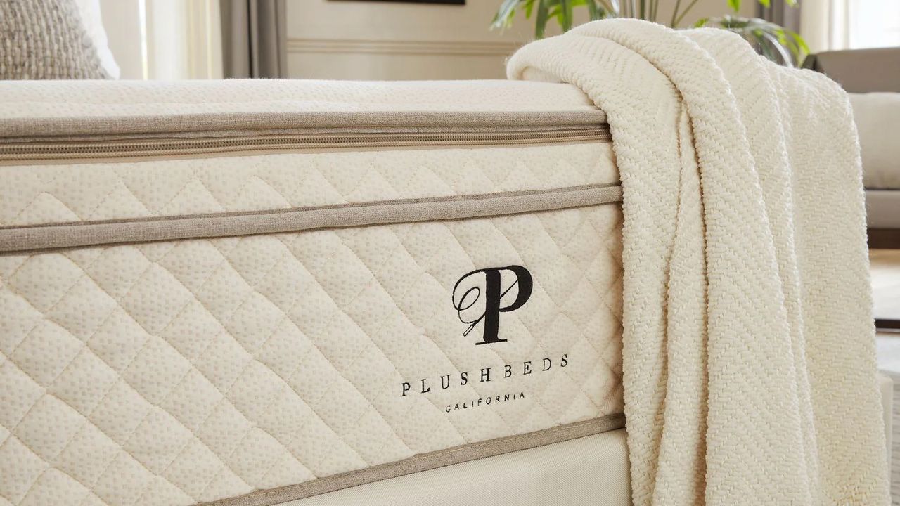 A closeup of the PlushBeds logo on the foot of the PlushBeds Botanical Bliss Mattress, with bedding draped over the end of the bed