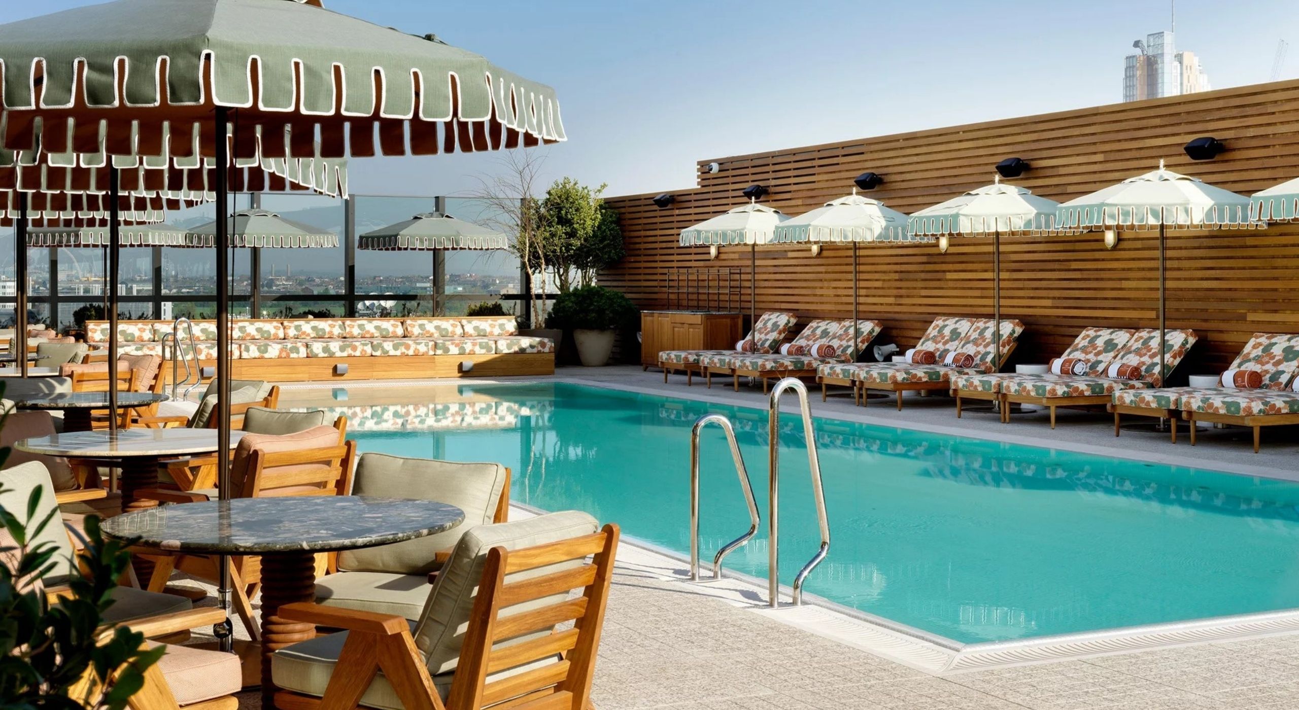 A beautifully decorated outdoor pool area, with sage green and wood furniture, round black marble tables, pale mint green parasols, and a wooden wall.