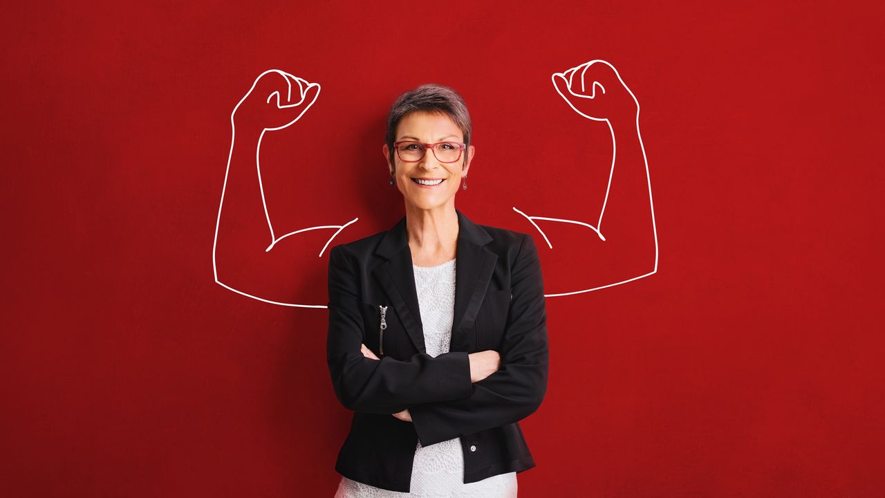 A woman stands against a red background with her arms crossed. Cartoon arms with big muscles are drawn on the red background on either side of her.