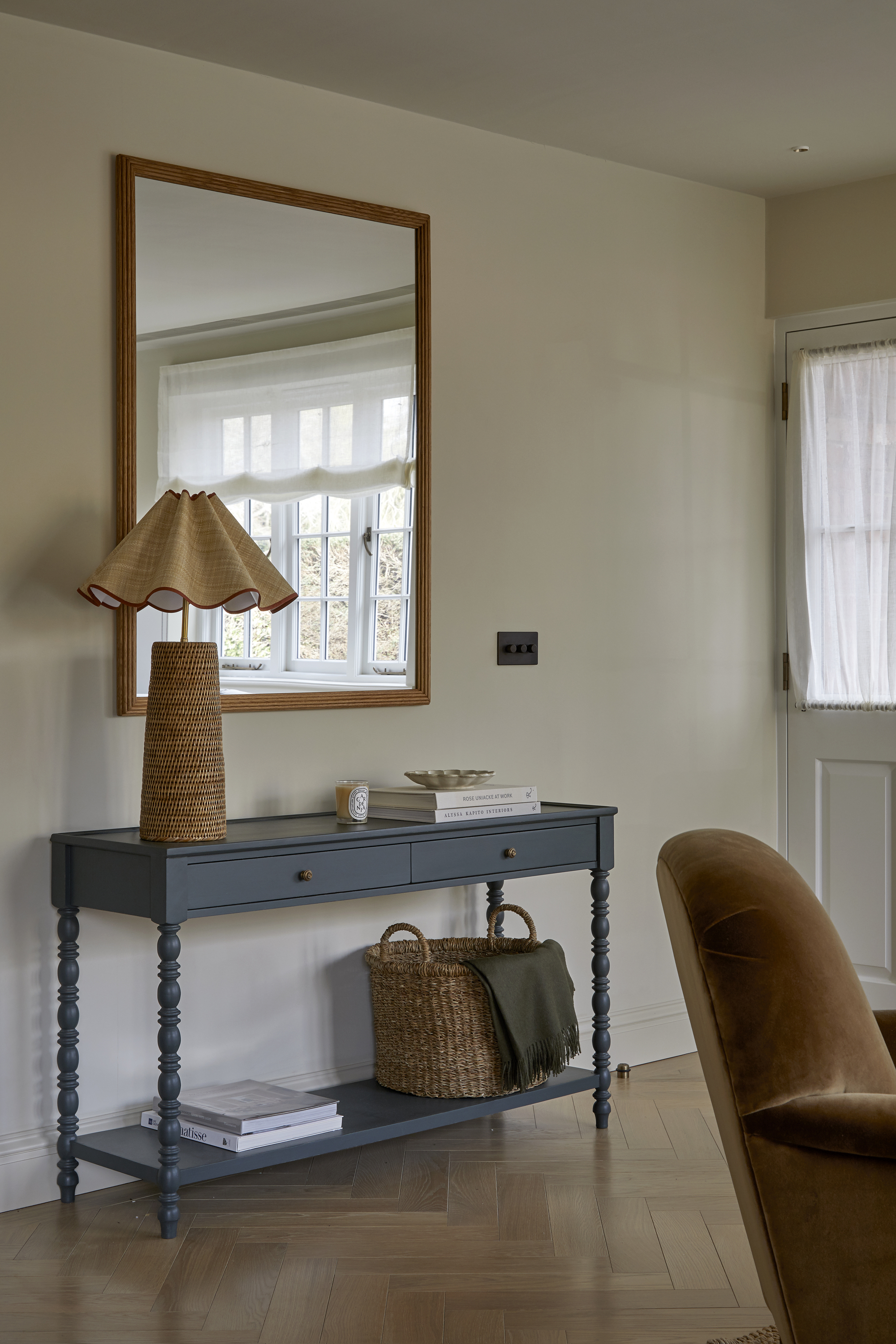 Image of a minimalist entryway that has a blue bobbin console table with a wicker lamp and basket on it. There is a mirror on the wall above the table and the walls are painted beige.