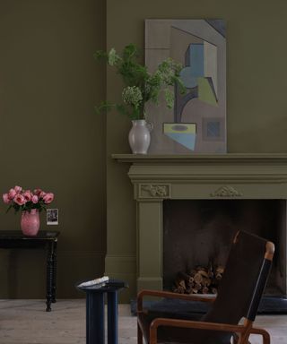 dark living room with olive green walls, fireplace and vase of foliage and artwork on mantel, black armchair in foreground