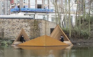 The floating platform placed on the river is constructed out of wood and is in the shape of three triangles that are connected. Two people are sitting on the platworm.