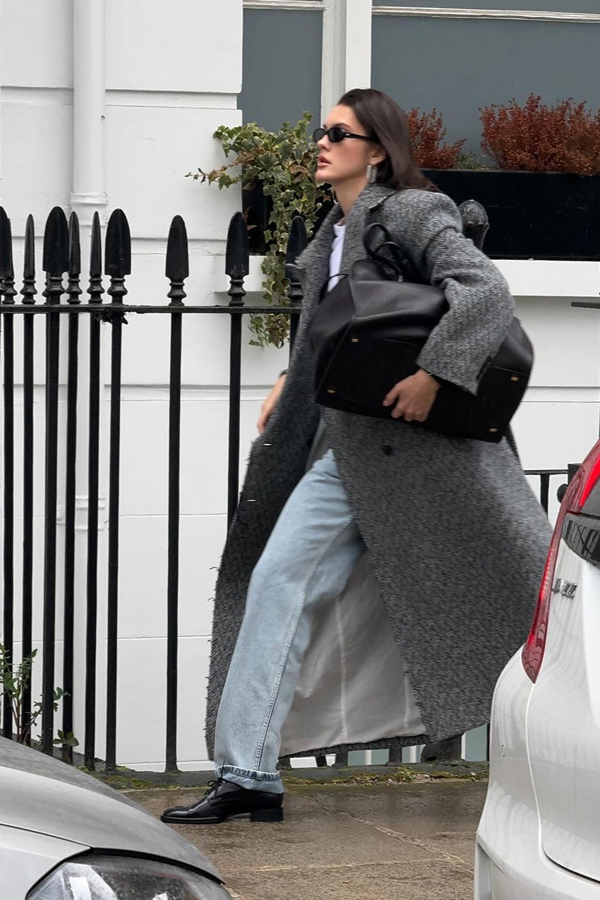 A woman walking down the sidewalk in London wearing black oval sunglasses, silver hoops, a long herringbone black and gray coat over a white T-shirt and light wash baggy jeans styled with black oxfords and a black bowler bag.